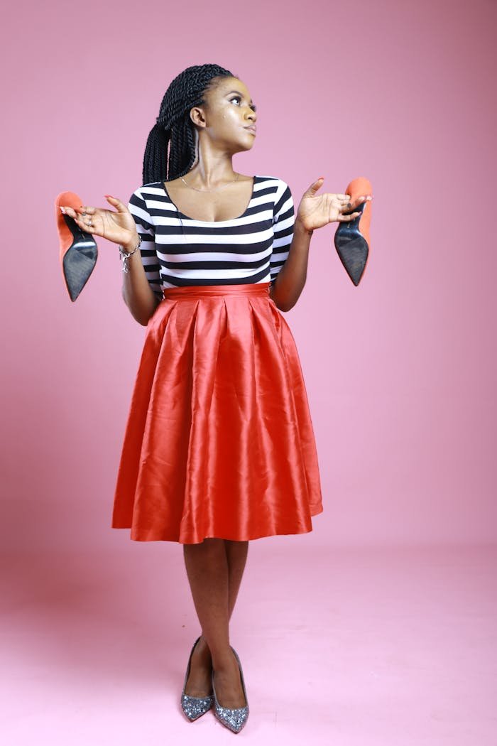 Full body of serious African American female in trendy skirt standing on pink background with crossed legs and pair of heels in hands